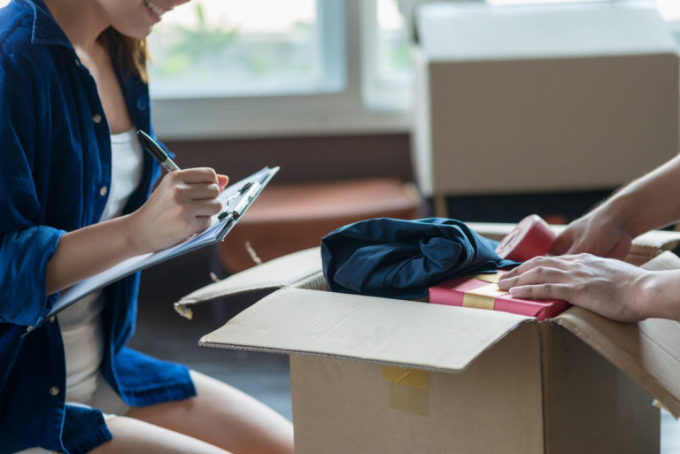 Woman kneeling next to full moving box, writing on a clipboard.