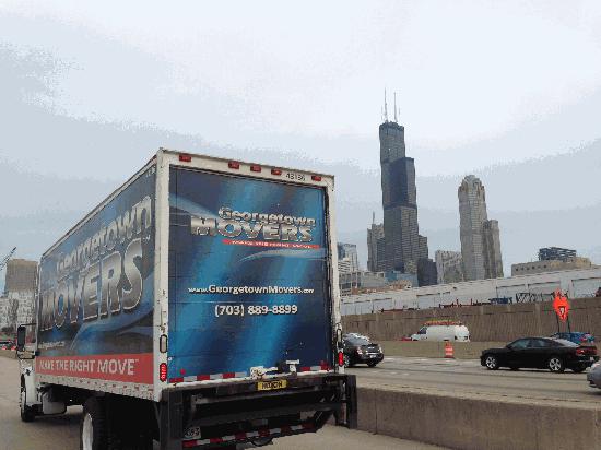Moving truck on side of road with city skyline in the background.